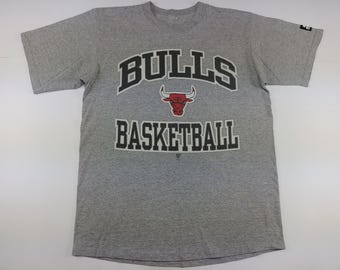 May include: Gray Bulls Basketball t-shirt. The shirt features the word "BULLS" in black block letters above a red bull logo, and the word "BASKETBALL" in black block letters below. The shirt is short-sleeved.