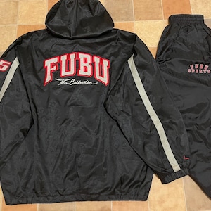 Può includere: Tuta Fubu nera con giacca con cappuccio e pantaloni abbinati. La giacca presenta il logo Fubu in rosso e bianco sul retro, con una striscia bianca sulle maniche. I pantaloni hanno il logo Fubu Sports.