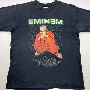 以下が含まれることがあります： 緑色の文字で「EMINEM」と白くプリントされた黒いTシャツ。シャツには、赤いジャンプスーツと白いスニーカーを履いて地面に座っているエミネムの写真がプリントされています。