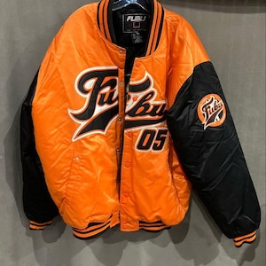 Può includere: Giacca bomber in raso arancione e nero con la scritta "FUBU" in bianco e nero sul davanti. Il numero "05" è sotto la scritta. Le maniche sono nere e la giacca ha colletto e polsini a righe nere e arancioni.