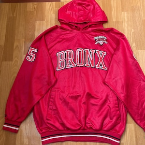 以下が含まれることがあります： 胸に白と赤の大きな文字で「BRONX」と書かれた赤いフード付きスウェットシャツ。左袖には数字の「5」が入っています。ドローストリングフードとフロントポケットが付いています。