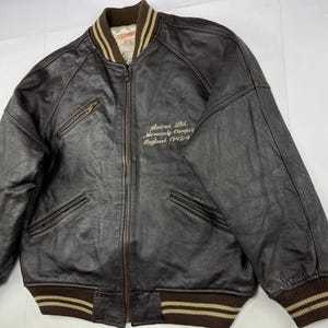 Pu&ograve; includere: Giacca bomber in pelle marrone con chiusura a zip, due tasche frontali e colletto, polsini e orlo a coste. La giacca presenta la scritta "Avirex Ltd. Normandy Camp England 1942-4" ricamata sul petto.