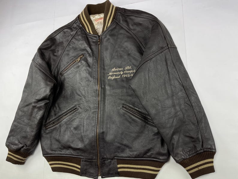 Pu&ograve; includere: Giacca bomber in pelle marrone con chiusura a zip, due tasche frontali e colletto, polsini e orlo a coste. La giacca presenta la scritta "Avirex Ltd. Normandy Camp England 1942-4" ricamata sul petto.