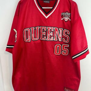 May include: Red baseball jersey featuring "QUEENS 05" in white and black lettering. The jersey has a V-neck with white and black trim, short sleeves, and a "FUBU ATHLET 1992" patch. The jersey is hanging on a white hanger.