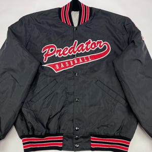 Può includere: Giacca da baseball in satin nero con bordini a righe rosse e bianche. La giacca ha una chiusura a bottoni e presenta il testo "Predator Baseball" in ricamo rosso e bianco.