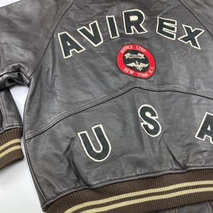 Pu&ograve; includere: Giacca bomber in pelle marrone con la scritta "AVIREX" in lettere bianche sul retro. Una toppa circolare con un'emblema alato e le parole "AVIREX LTD NEW YORK" &egrave; centrata sotto le lettere. Viene visualizzata anche la parola "USA".