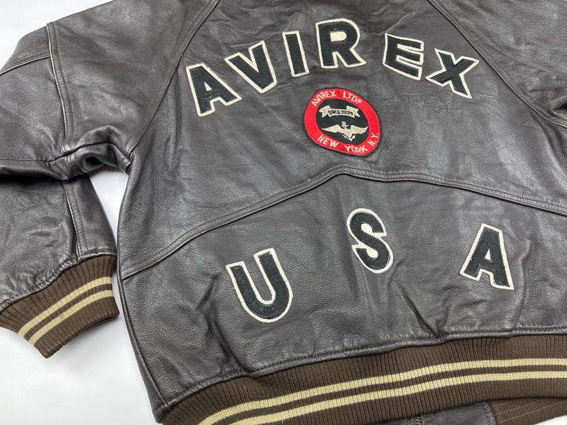 Pu&ograve; includere: Giacca bomber in pelle marrone con la scritta "AVIREX" in lettere bianche sul retro. Una toppa circolare con un'emblema alato e le parole "AVIREX LTD NEW YORK" &egrave; centrata sotto le lettere. Viene visualizzata anche la parola "USA".