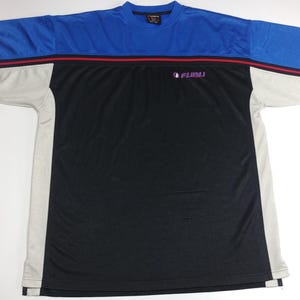 May include: A blue, black, and grey short-sleeved t-shirt featuring the brand name "FUBU" in purple. The shirt has a colour block design with a blue upper section, a black lower section, and grey side panels. A red stripe accents the blue section.