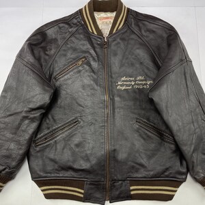 Pu&ograve; includere: Giacca bomber in pelle marrone con chiusura a zip, due tasche frontali e polsini e orlo a coste. La giacca presenta la scritta ricamata "Avirex Ltd. Normandy Campaign England 1942-43" sul petto.