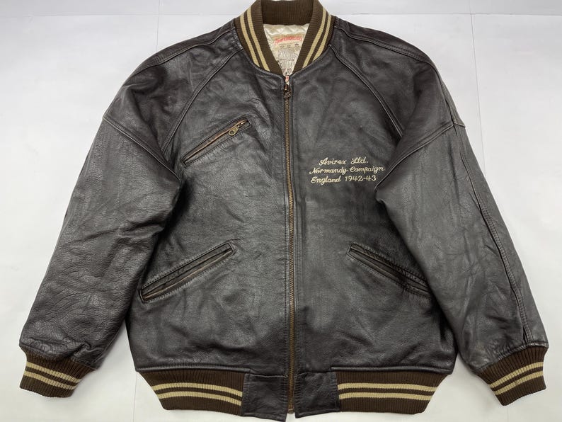 Pu&ograve; includere: Giacca bomber in pelle marrone con chiusura a zip, due tasche frontali e polsini e orlo a coste. La giacca presenta la scritta ricamata "Avirex Ltd. Normandy Campaign England 1942-43" sul petto.