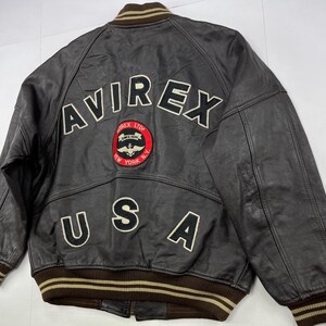 Pu&ograve; includere: Giacca bomber in pelle marrone con la scritta "AVIREX" in lettere bianche sul retro. Sotto c'&egrave; una toppa circolare con le parole "AVIREX LTD SINCE 1975 NEW YORK N.Y." Anche la parola "USA" &egrave; sulla giacca.
