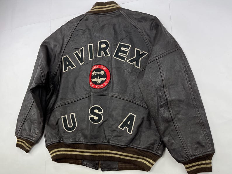 Pu&ograve; includere: Giacca bomber in pelle marrone con la scritta "AVIREX" in lettere bianche sul retro. Sotto c'&egrave; una toppa circolare con le parole "AVIREX LTD SINCE 1975 NEW YORK N.Y." Anche la parola "USA" &egrave; sulla giacca.