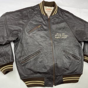 Pu&ograve; includere: Giacca bomber in pelle marrone scuro con chiusura a zip, due tasche frontali e polsini e orlo a coste. La giacca ha un colletto e polsini a righe marroni e beige. Il testo "Avirex Ltd. Normandy Campaign England 1942-43" &egrave; ricamato sul petto.
