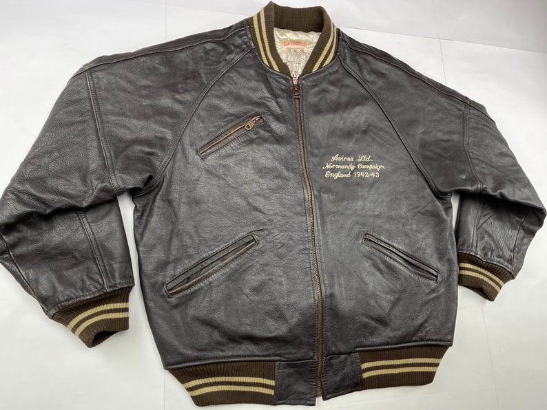 Pu&ograve; includere: Giacca bomber in pelle marrone scuro con chiusura a zip, due tasche frontali e polsini e orlo a coste. La giacca ha un colletto e polsini a righe marroni e beige. Il testo "Avirex Ltd. Normandy Campaign England 1942-43" &egrave; ricamato sul petto.