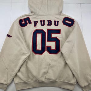 May include: Beige hoodie with a hood and long sleeves. The back features the word "FUBU" and the number "05" in navy blue with red trim. The number "5" and "0" are on the shoulders. The hoodie has a small "FUBU" logo on the left sleeve.