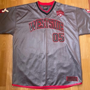 Peut inclure: Maillot de baseball gris avec des détails rouges et noirs. Le maillot affiche le mot "WESTSIDE" en rouge avec un contour noir, le numéro "05" et un logo sur la poitrine. Les manches ont des rayures rouges.