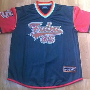 May include: A black V-neck jersey with red sleeves featuring orange and black stripes. The front displays the text Fubu and 05 in orange and white. The number 05 appears on the right sleeve. A rectangular patch with text is located at the bottom right.