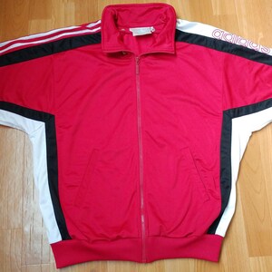 May include: A red zip-up track jacket with white and black accents. The jacket features white stripes on the sleeves and the Adidas logo in white on the upper sleeve. The jacket has a high collar and two front pockets.