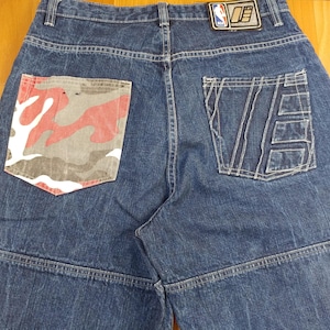 May include: Dark blue denim jeans featuring a high waist and a rectangular NBA logo patch on the back waistband. The left back pocket displays a red, white, and gray camouflage pattern, while the right back pocket features stitched geometric lines.
