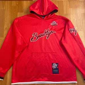 May include: A red hoodie with a hood and a front pocket. The word "Brooklyn" is written in white script across the chest. The sleeves have black and white trim at the cuffs. The letters "NY" are on the right sleeve.