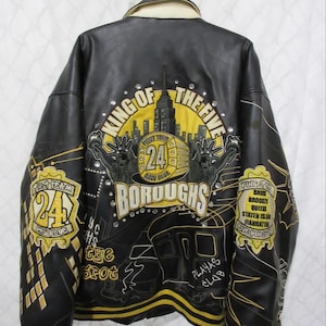 Könnte beinhalten: Schwarze und gelbe Collegejacke mit dem Text "King of the Five Boroughs" und "24 Raw Blue". Die Jacke zeigt ein Stadtbilddesign und die Freiheitsstatue. Die Jacke hat einen gelb-schwarzen Streifen.