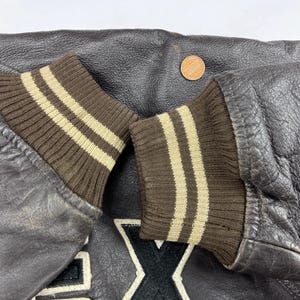 Pu&ograve; includere: Primo piano di una giacca di pelle marrone scuro con polsini a coste marroni. I polsini hanno due strisce beige chiaro. Una moneta &egrave; visibile sulla pelle.