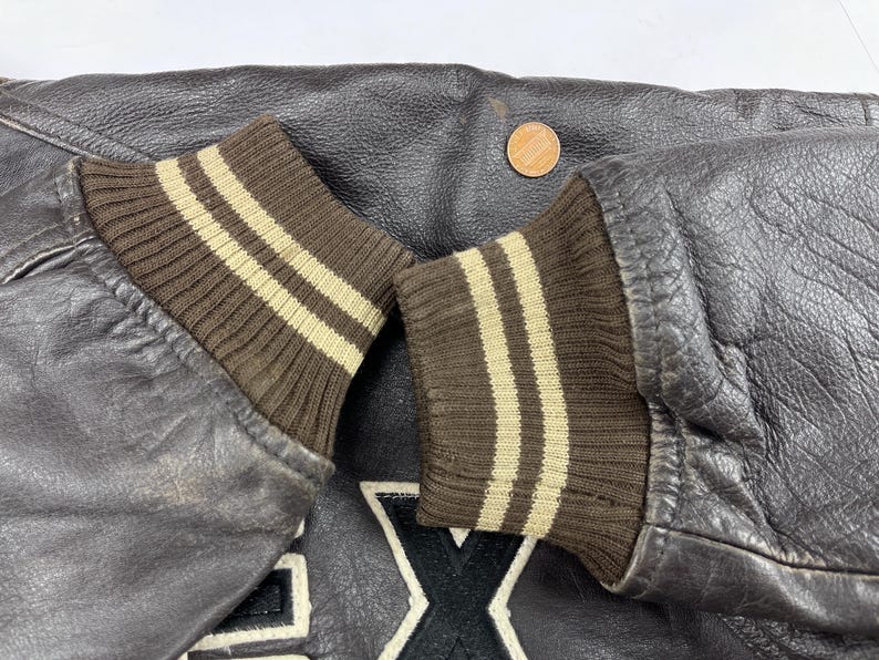 Pu&ograve; includere: Primo piano di una giacca di pelle marrone scuro con polsini a coste marroni. I polsini hanno due strisce beige chiaro. Una moneta &egrave; visibile sulla pelle.