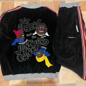 May include: Black velour tracksuit with a jacket and trousers. The jacket has red and white stripes on the sleeves and the phrase "FAT ALBERT and the JUNKYARD GANG!" on the back. The trousers have red and white stripes on the side.