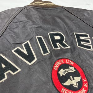 Pu&ograve; includere: Primo piano di una giacca di pelle marrone con la scritta "AVIREX" a grandi lettere bianche. &Egrave; visibile anche una toppa circolare con le scritte "AVIREX LTD" e "NEW YORK". La giacca ha un colletto a righe marrone e beige.
