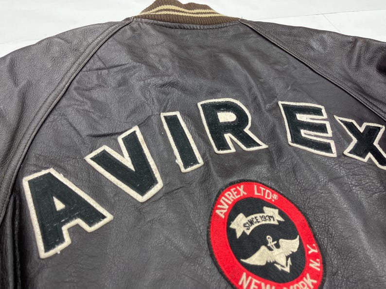 Pu&ograve; includere: Primo piano di una giacca di pelle marrone con la scritta "AVIREX" a grandi lettere bianche. &Egrave; visibile anche una toppa circolare con le scritte "AVIREX LTD" e "NEW YORK". La giacca ha un colletto a righe marrone e beige.