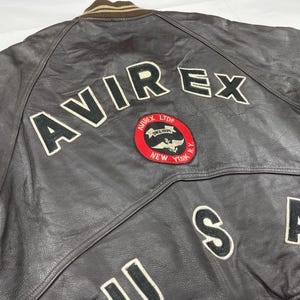 Pu&ograve; includere: Giacca bomber in pelle marrone con la scritta "AVIREX" in grandi lettere bianche bordate di nero sul retro. Una toppa circolare con le parole "AVIREX LTD NEW YORK" &egrave; centrata sotto il nome. Sono visibili anche le lettere "USA".