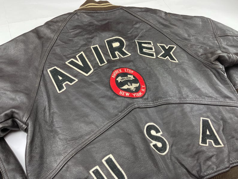 Pu&ograve; includere: Giacca bomber in pelle marrone con la scritta "AVIREX" in grandi lettere bianche bordate di nero sul retro. Una toppa circolare con le parole "AVIREX LTD NEW YORK" &egrave; centrata sotto il nome. Sono visibili anche le lettere "USA".