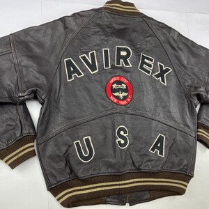 Pu&ograve; includere: Giacca bomber in pelle marrone con la scritta "AVIREX" in lettere bianche sulla schiena. Sotto la scritta "AVIREX" c'&egrave; una toppa con un cerchio rosso e un'emblema alato. La parola "USA" &egrave; presente anche sul retro.