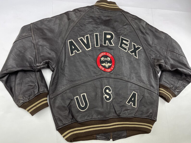 Pu&ograve; includere: Giacca bomber in pelle marrone con la scritta "AVIREX" in lettere bianche sulla schiena. Sotto la scritta "AVIREX" c'&egrave; una toppa con un cerchio rosso e un'emblema alato. La parola "USA" &egrave; presente anche sul retro.