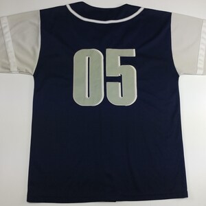 May include: Navy blue baseball jersey with white and grey accents. The jersey features the number "05" in a light grey colour on the back and on the sleeves. The collar and sleeve cuffs are white.