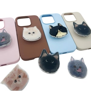 May include: A collection of phone cases in various colors, each featuring a different cat face design. The cases are in shades of pink, brown, blue, and beige. The cat faces are in various colors and styles, including black, white, and gray.