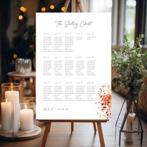 May include: A white seating chart on an easel with the words "The Seating Chart" in elegant script. The chart lists names under table numbers, with floral accents in the corner. The date "04 06 24" is at the bottom.