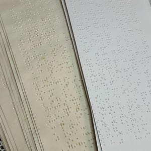 May include: A stack of Braille pages, some aged with a yellowed hue, others bright white. The raised dots form text, offering a tactile reading experience. The pages are stacked, suggesting a book or document.