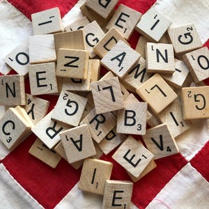May include: A collection of wooden Scrabble tiles scattered on a red and white quilted surface. The tiles feature black letters and point values. The background is a patchwork of red and white fabric.