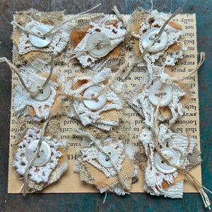 May include: Handmade fabric embellishments with white crochet lace, tan fabric, and pearl buttons. Each piece is attached with twine. The background is a vintage book page.