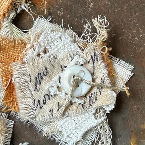 May include: A handmade textile art piece featuring a white button, stitched onto a beige fabric square with handwritten text. The piece incorporates frayed edges, lace, and textured burlap in shades of beige and orange.