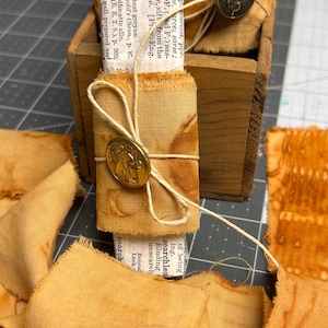 May include: A bundle of mustard-yellow fabric with frayed edges wrapped around a piece of printed dictionary paper. The bundle is secured with thin twine and a round, metallic gold-toned button featuring an embossed design on the front.