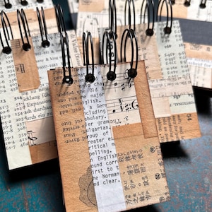 May include: A collection of small, rectangular notebooks bound with black wire spirals. The covers feature a collage of vintage-style paper, including printed English and Japanese text, musical notation, and numerical tables in beige and black.