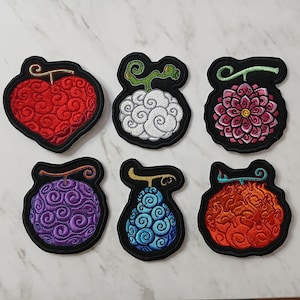 May include: Set of six embroidered patches featuring colorful, stylized fruit designs. Each patch has a black border and depicts a unique fruit with intricate details in red, white, pink, purple, blue, and orange. The patches are likely for clothing or accessories.