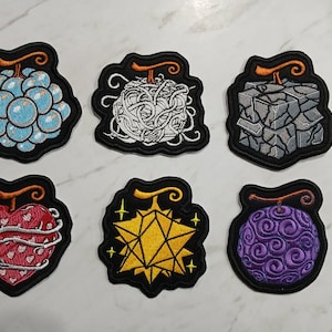 Puede incluir: Seis parches bordados con varios diseños de frutas. Los parches tienen un borde negro y un tallo. Las frutas incluyen un grupo de burbujas azules, una bola de hilos blancos, una piedra gris, un corazón rojo, una estrella amarilla y un remolino púrpura.
