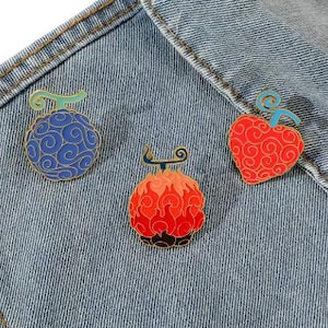 May include: Three enamel pins shaped like stylised fruits with swirling patterns. One pin is blue with a light green stem, one is red with a blue stem, and the centre pin features a gradient of orange and red flames with a dark stem.
