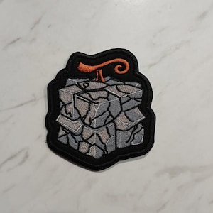 May include: Embroidered patch with a grey, stone-like design, a black border, and an orange swirl. The patch has a detailed, textured appearance, suitable for application on clothing or accessories.