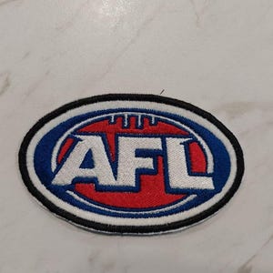 May include: A white, blue, and red embroidered patch with the letters "AFL" inside a circle. The patch is on a white background.