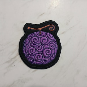 May include: Embroidered patch featuring a purple, swirling fruit design with an orange stem, set against a black background. The patch has a unique shape and is likely for decorative purposes, possibly for clothing or accessories.