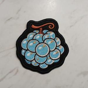 May include: Embroidered patch depicting a cluster of light blue orbs with white highlights, resembling a fruit, set against a black border. A brown stem and tendril are at the top. Suitable for clothing or accessories.